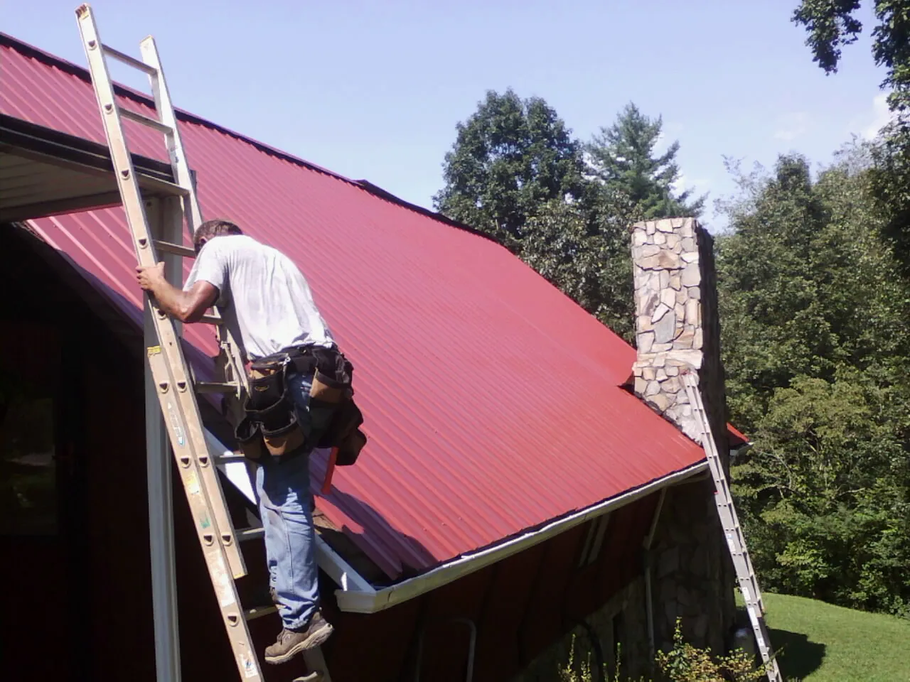 Roofer on a ladder beside a red metal roof and stone chimney — Soffit & Fascia Repair by Roofing Brothers in Burns